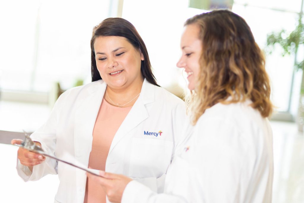 Two Mercy caregivers in lab coats looking at a clipboard.