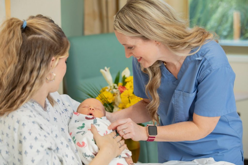 An image of a new mother holding her swaddled newborn while a Mercy nurse smiles and gently touches the mother and baby.