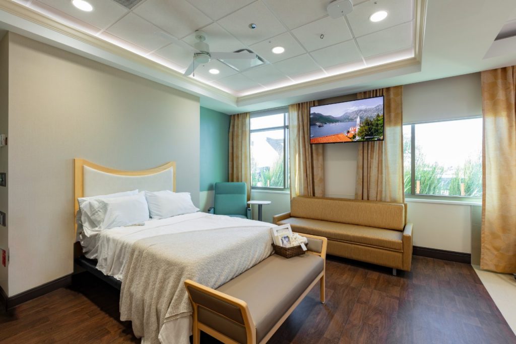 A birthing suite in the Love Family Women's Center at Mercy Hospital Oklahoma City. It shows a queen-sized, home-style bed used to mimic a home birth setting in the hospital. There is recessed lighting and crown-wood molding on the ceiling, a large sleeping couch and a rocking chair behind the bed, plus a large TV, and two large windows with blinds and curtains.