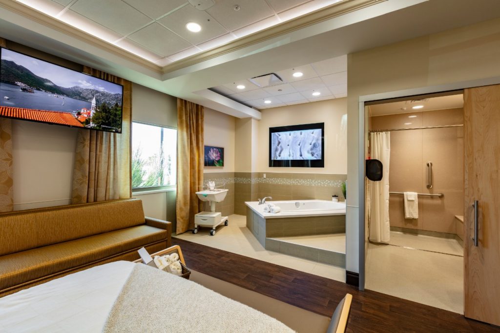 A birthing suite in the Love Family Women's Center at Mercy Hospital Oklahoma City. It shows a queen-sized, home-style bed used to mimic a home birth setting in the hospital. There is recessed lighting and crown-wood molding on the ceiling, a large sleeping couch and a rocking chair behind the bed, plus a large TV, and two large windows with blinds and curtains.