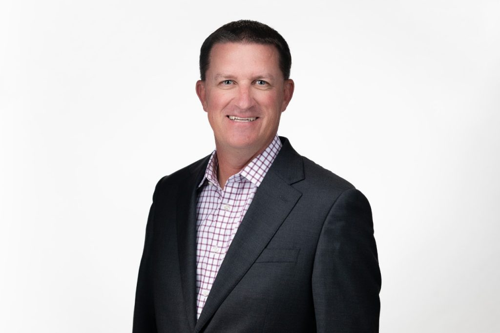 A head shot of Dustin Yowell, smiling in a checkered Oxford-collared shirt and a dark sport coat.