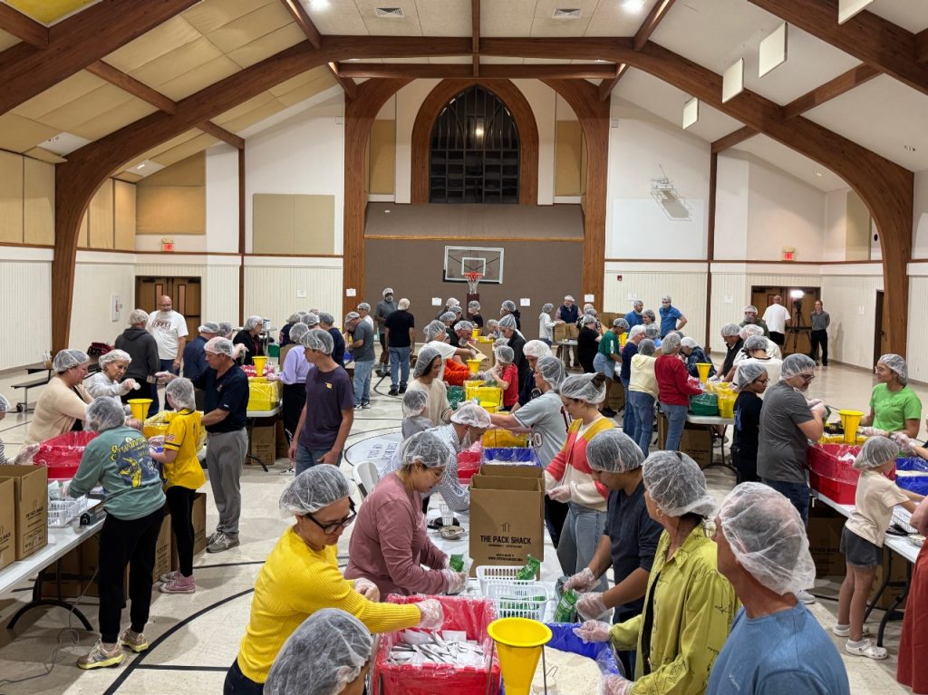 Dozens of volunteers in hair nets stand around tables to make assembly lines for a food donation drive in a church school gym.
