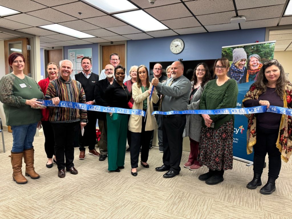 A group of about 20 people standing in an office area cutting a blue ribbon with large ceremonial scissors.