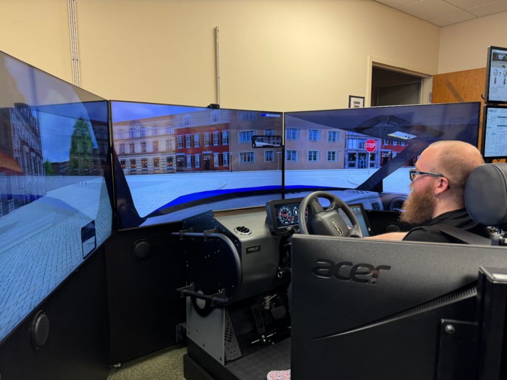 A photo from behind and to the left of a Mercy caregiver seated at an immersive driving simulator for rehabilitation patients.