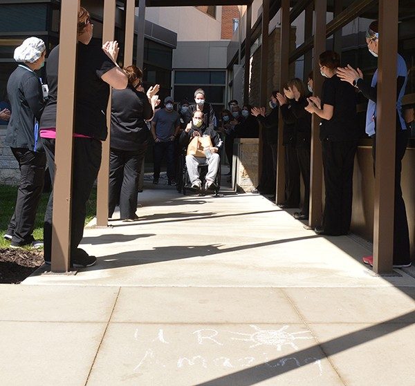 Patrick Linderer gives a thumbs up to cheering hospital co-workers as he is wheeled to his vehicle at the discharge canopy. The chalk art in the foreground could apply to him and all his caregivers.