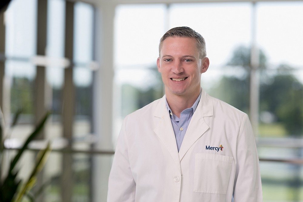 Dr. Caleb Norton poses smiling wearing a white lab coat with a Mercy logo