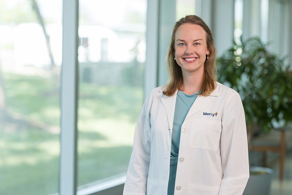 Dr. Becky Jadwisiak poses and smiles while wearing a white lab coat with a Mercy logo