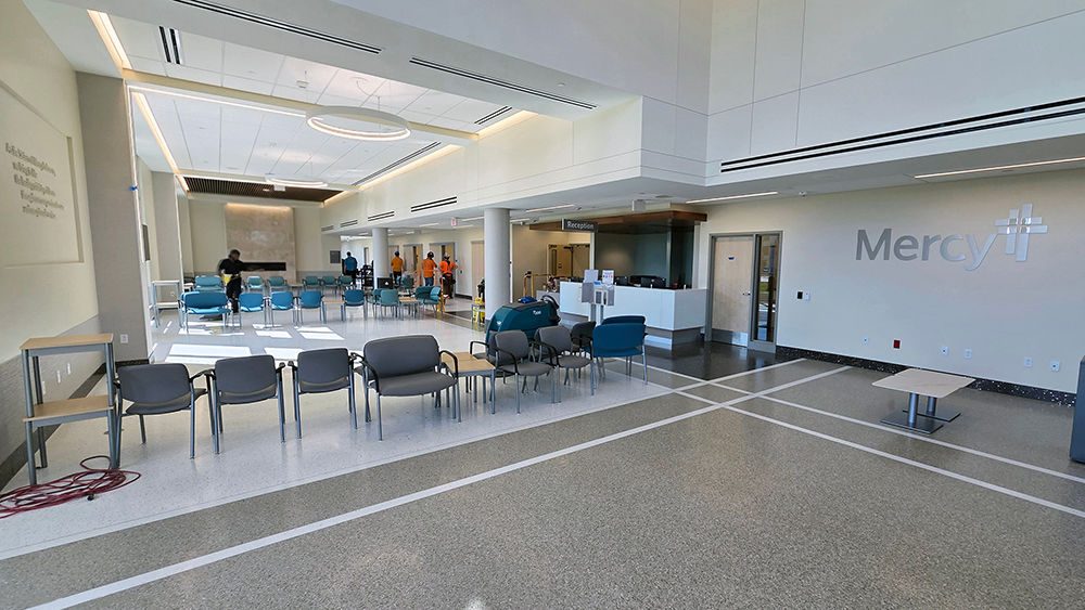 A large waiting room with chairs and tables is well lit by floor-to-ceiling windows with a fireplace on the back wall.