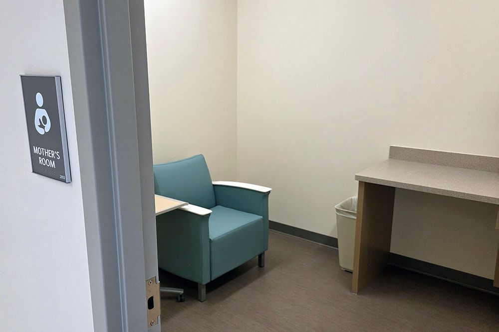 A green chair with a table next to it and a counter in a room. A sign reading "Mother's Room" hangs on the wall outside the room.